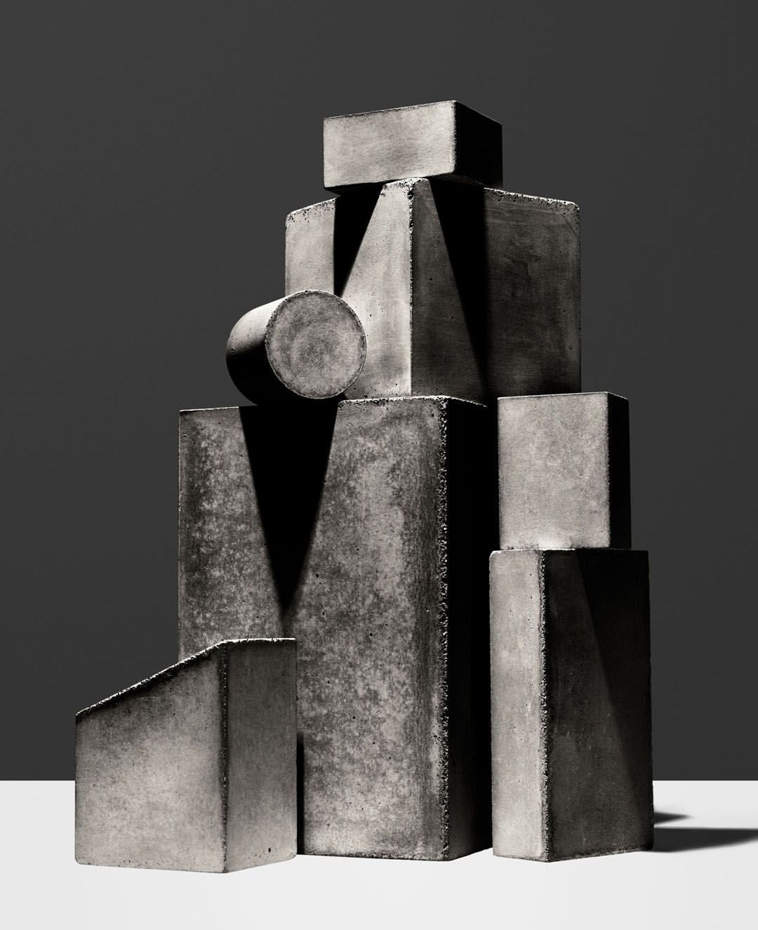 Monochromatic studio still life features a textured stacked geometric shapes, casting stark shadows on a gradient gray background.