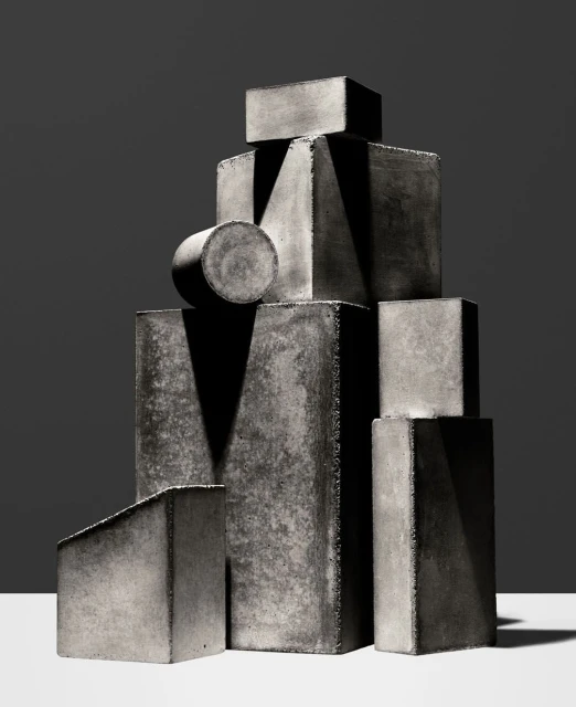 Monochromatic studio still life features a textured stacked geometric shapes, casting stark shadows on a gradient gray background.