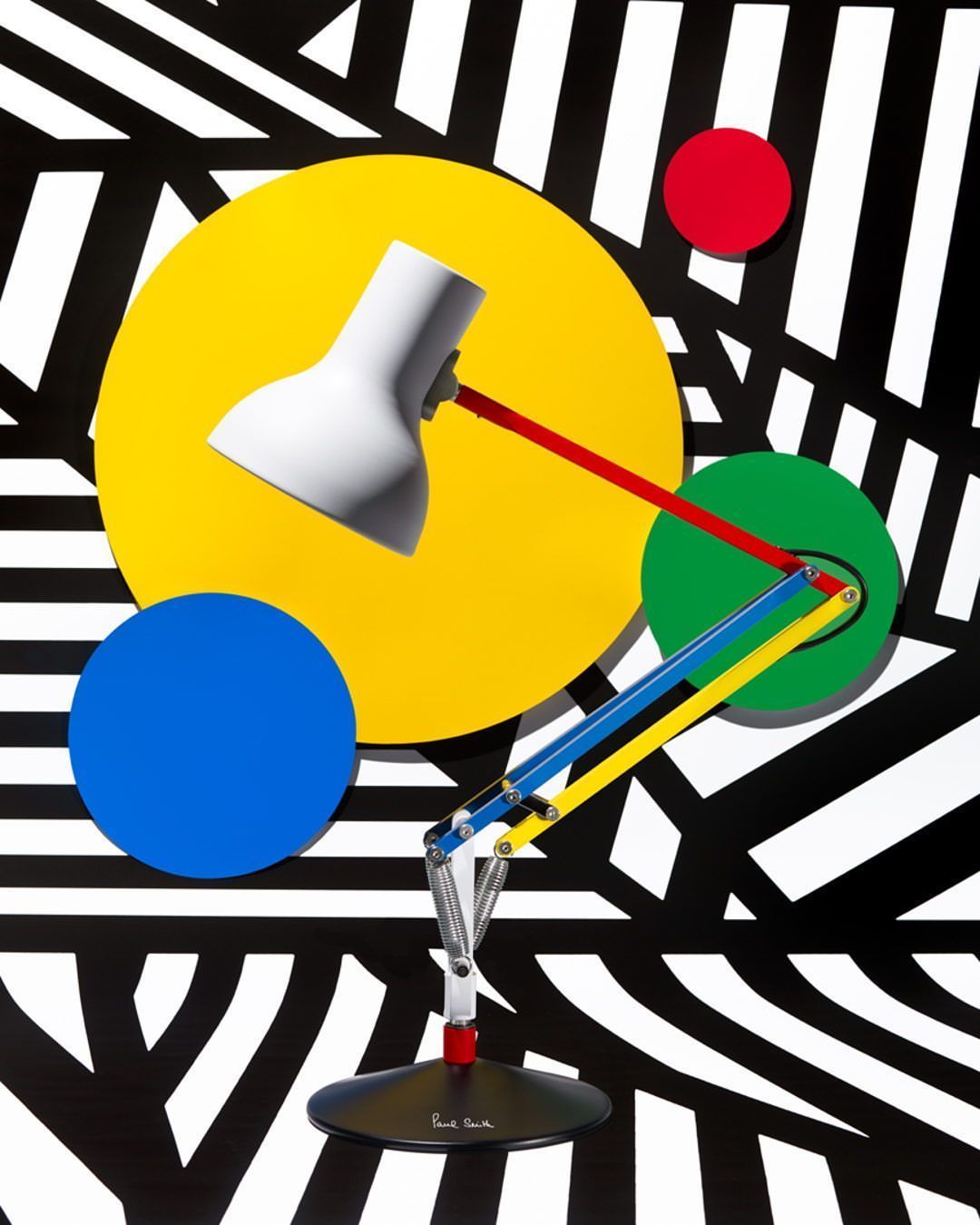 Abstract product photography featuring a white adjustable desk lamp with colorful circular backdrops, set against a black and white striped background.