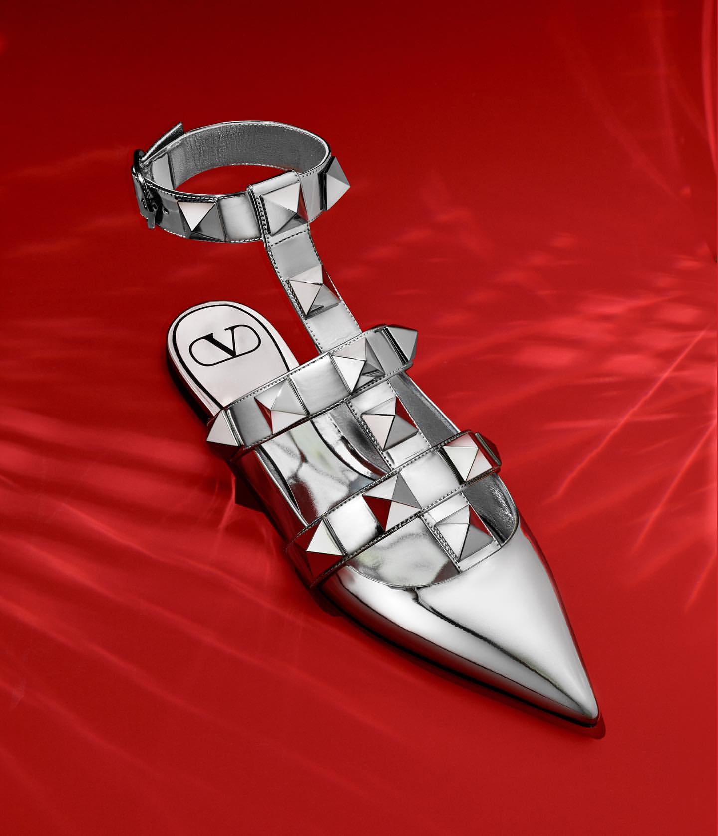 Silver Valentino Garavani studded sandal on a red silk surface, showing the pointed toe and ankle strap design. Fashion editorial.