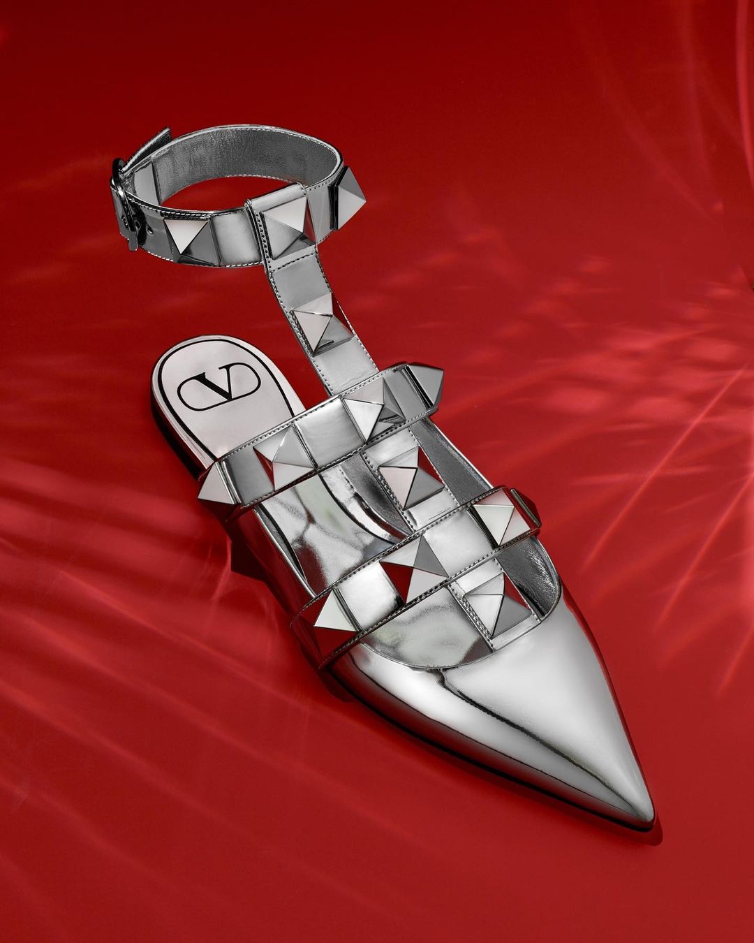 Silver Valentino Garavani studded sandal on a red satin background. The sandal has a pointed toe and an ankle strap with a buckle.