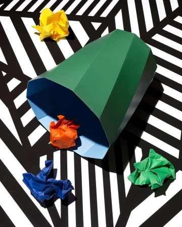 Eye-level shot of a green polygonal container, with crumpled paper balls of yellow, orange, blue, and green on a black-and-white striped surface.