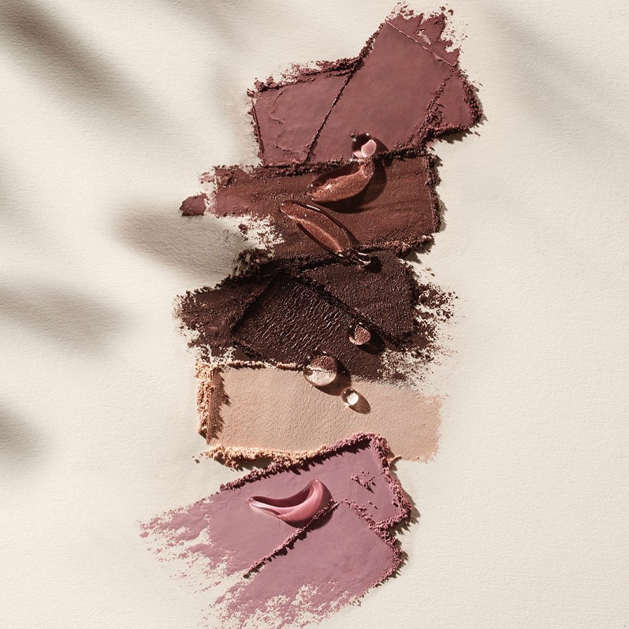 Close-up of swatches of brown and mauve eyeshadow and lipsticks, with water droplets, displayed on a textured white surface under shadow.
