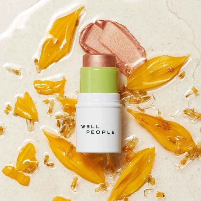 Well People makeup stick on a wet surface with yellow petals, in a beauty shot featuring a natural cosmetics product.