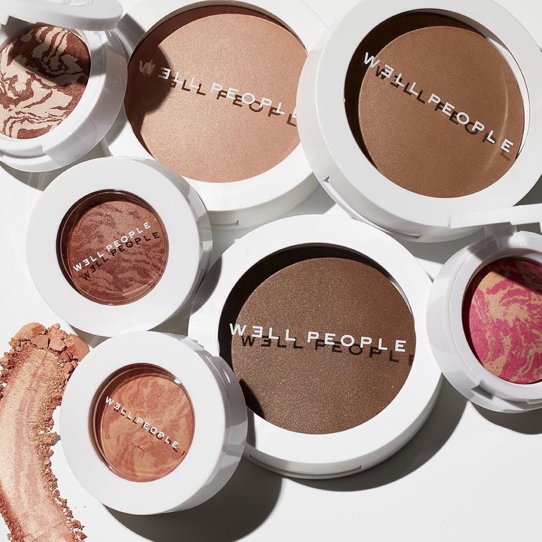 Cosmetic makeup products with bronzer and blushes from Well People are showcased with white compacts in a studio setting.