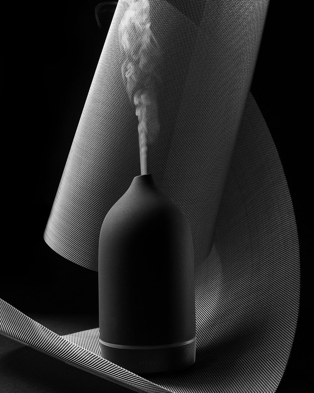 Minimalist black diffuser with vapor plume on dark backdrop with textured white surface in black and white studio product shot.