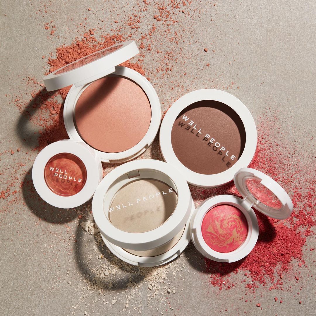 Five round compacts of various shades of blush and powder are arranged on a neutral surface with scattered powder.