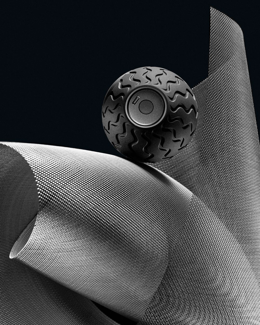 A textured ball with a speaker-like center sits atop a curved, dotted material against a dark background, creating a modern product still life.