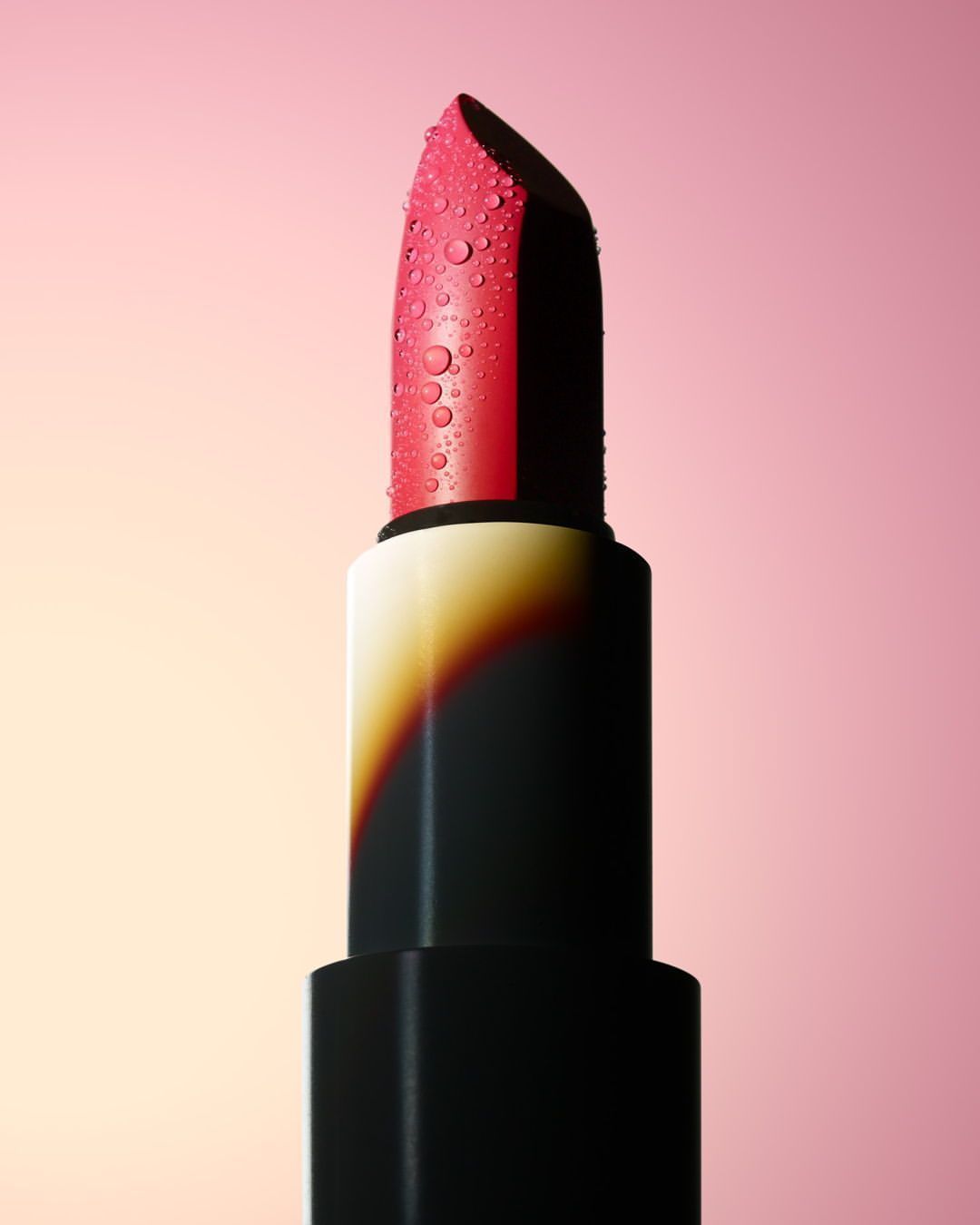 Close-up macro shot of a bright pink lipstick covered in tiny water droplets against a smooth gradient pink background.