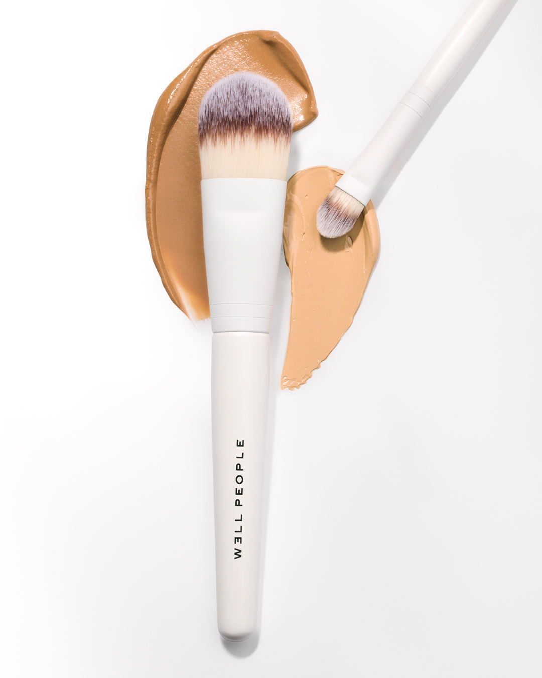 Close-up of foundation makeup brushes with white handles and synthetic bristles, displayed on a clean white surface with swipes of tan foundation.
