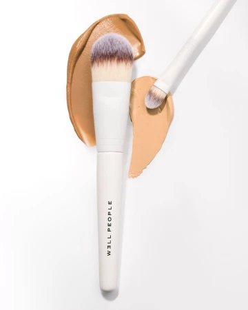 Close-up of foundation makeup brushes with white handles and synthetic bristles, displayed on a clean white surface with swipes of tan foundation.