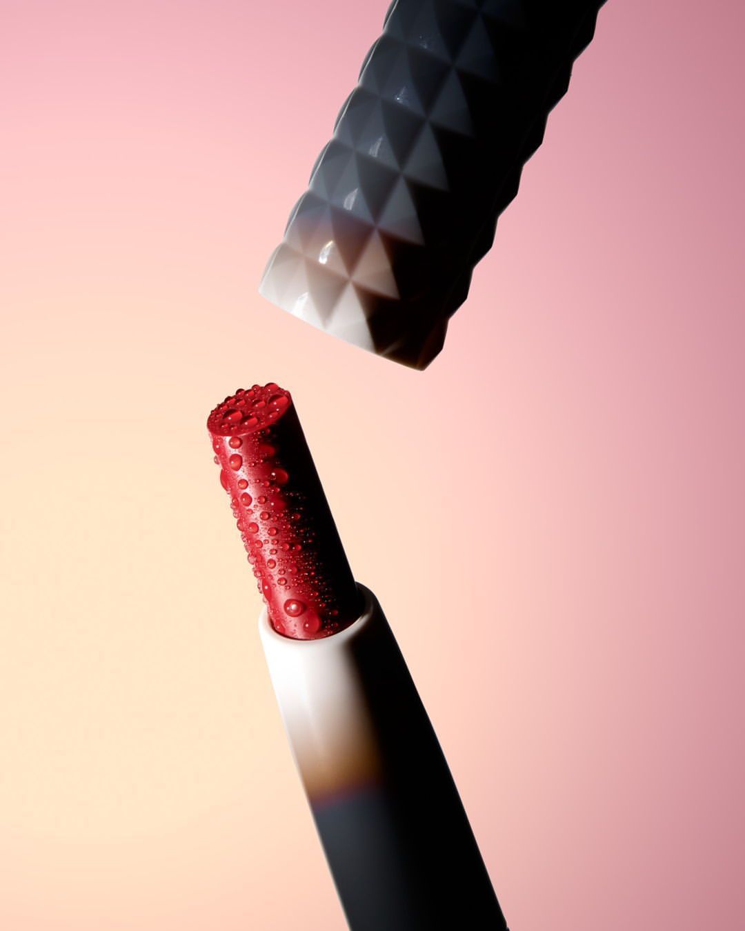 Close-up studio shot of red lipstick bullet with water droplets and its cap with a pink-to-yellow gradient background.