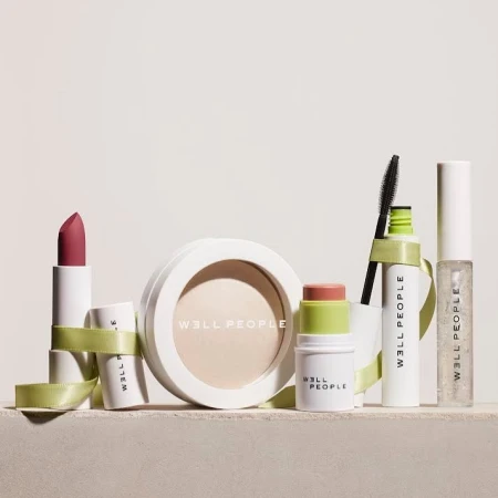 Well People beauty product assortment featuring lipstick, powder, blush stick, mascara, and lip gloss against a white backdrop.