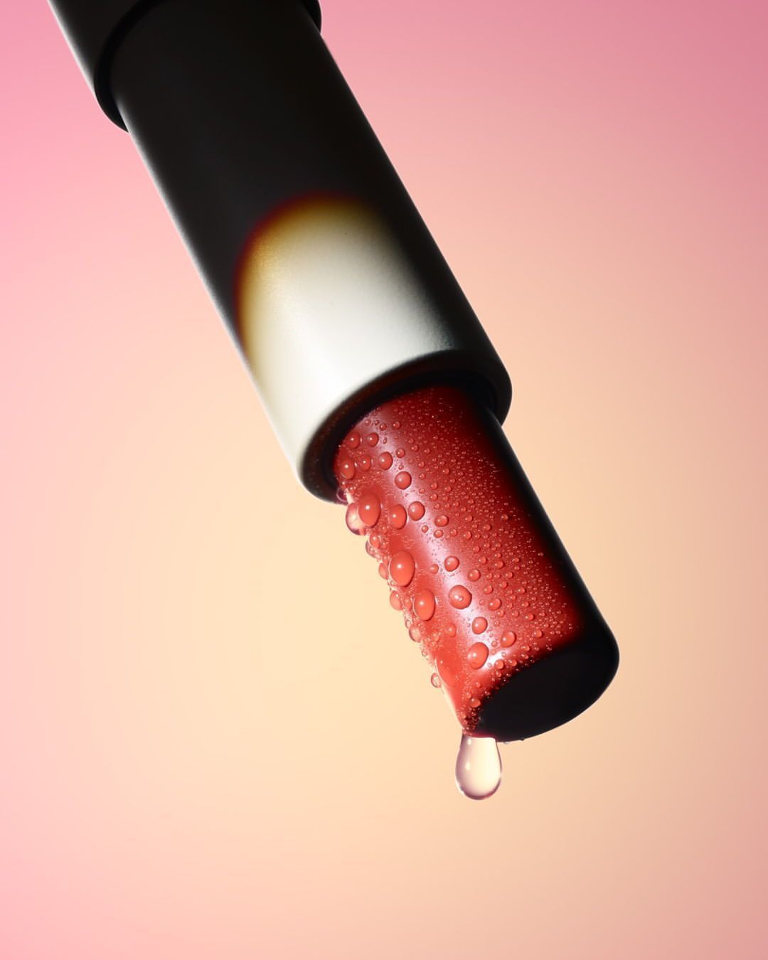 Close-up studio shot of red lipstick with water droplets on the tip and a single drop falling against a pink and yellow background.
