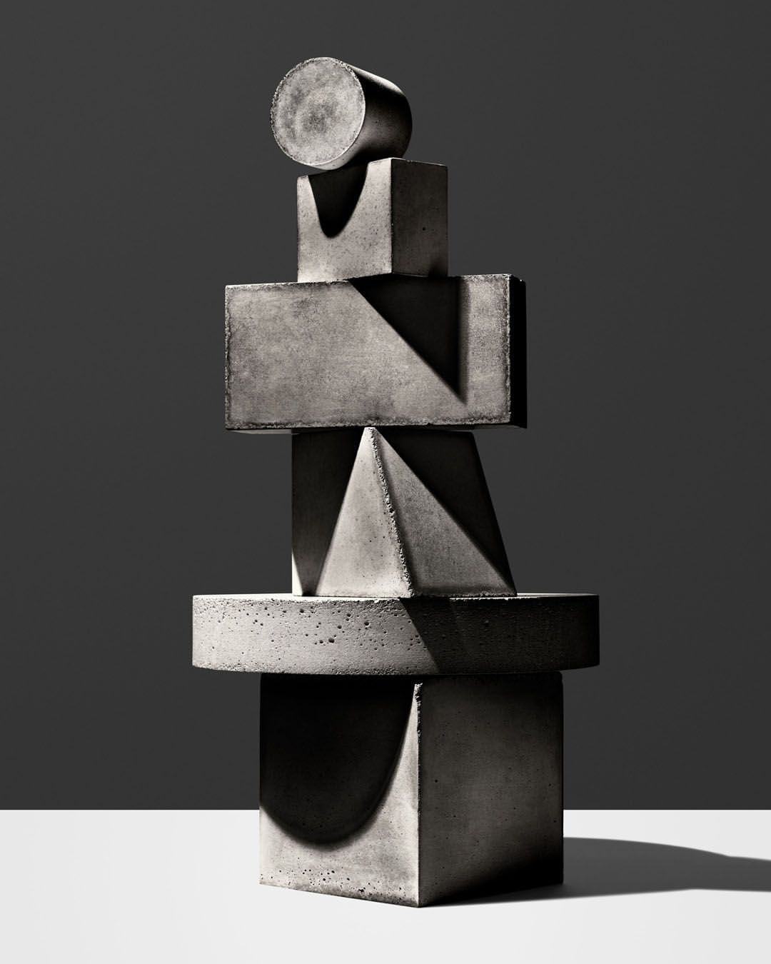 Still life of stacked geometric concrete shapes in studio setting, featuring cylinder, cubes, pyramid, and circle under direct lighting.