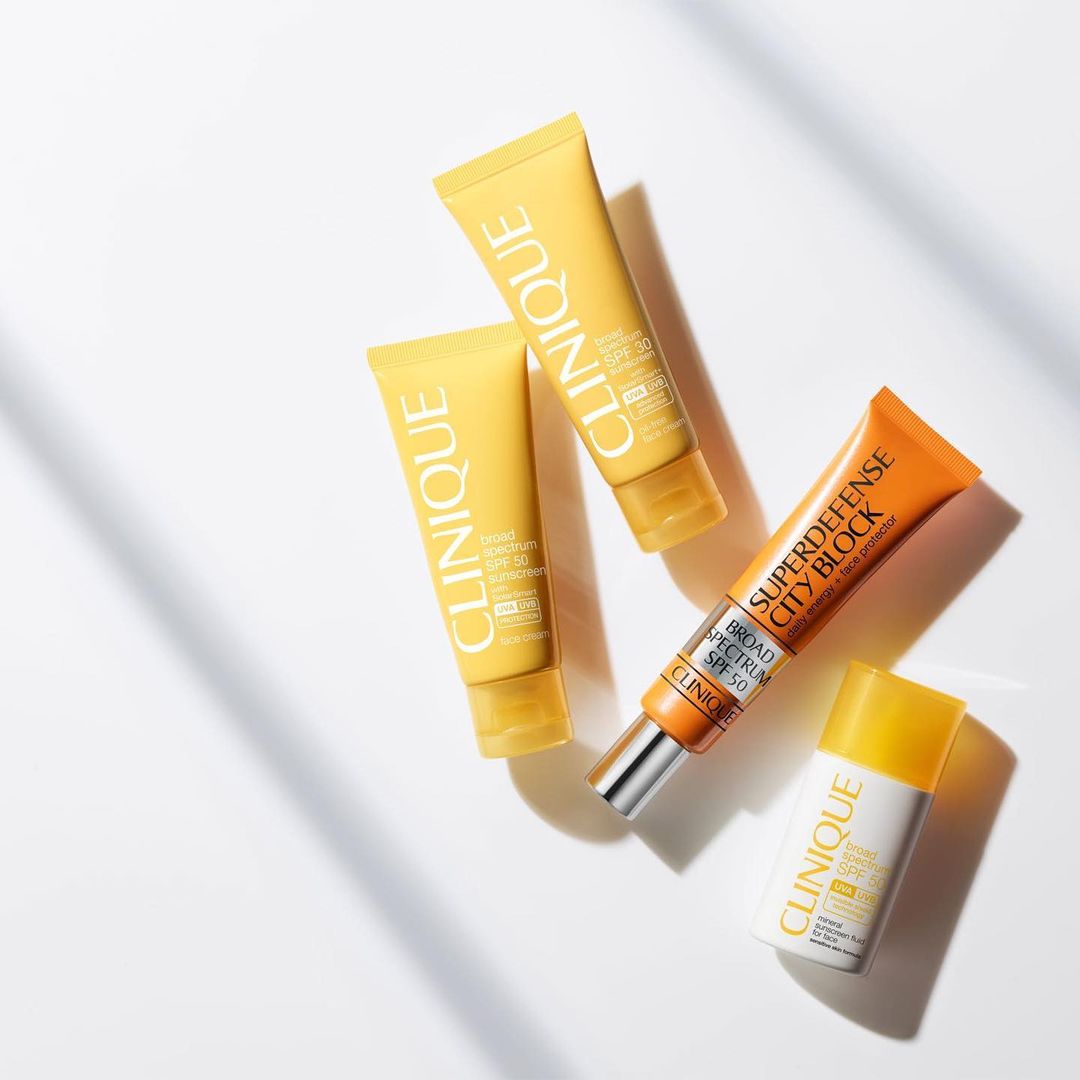 Four Clinique sunscreen tubes in yellow and orange hues are arranged on a white surface, casting soft shadows from the tubes.