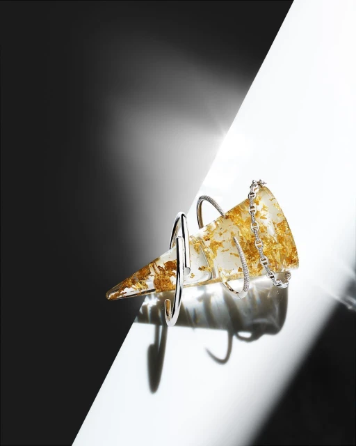 Still life shot of a translucent cone with gold inclusions and silver bracelets, set against a stark black and white background.