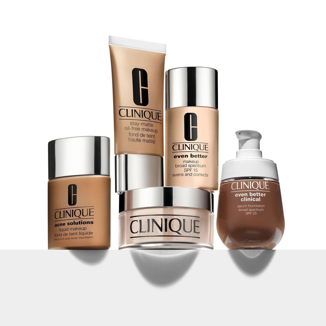 Several bottles and containers of Clinique makeup foundation in various tones are arranged on a white surface, studio lighting.
