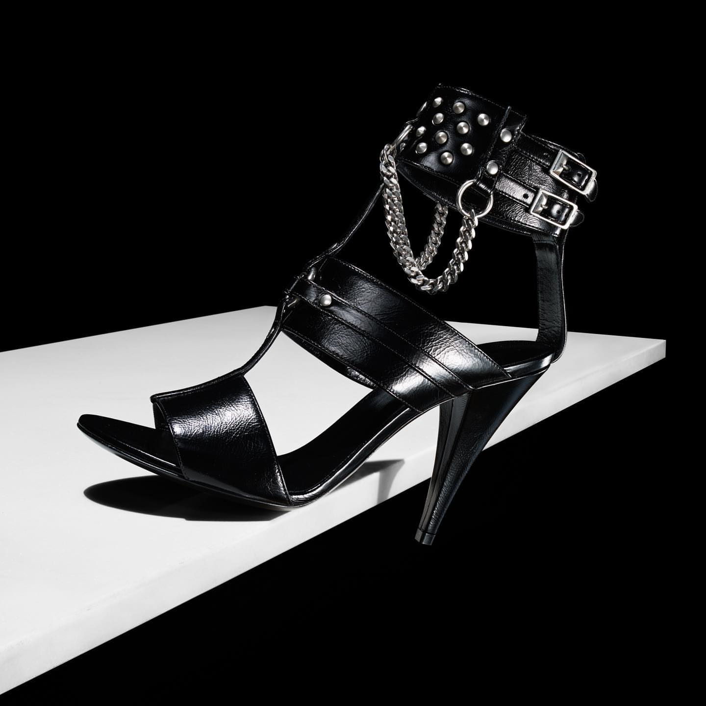 Black leather high-heel sandal with chain and studs accents on a white surface against a black background in a studio shot.