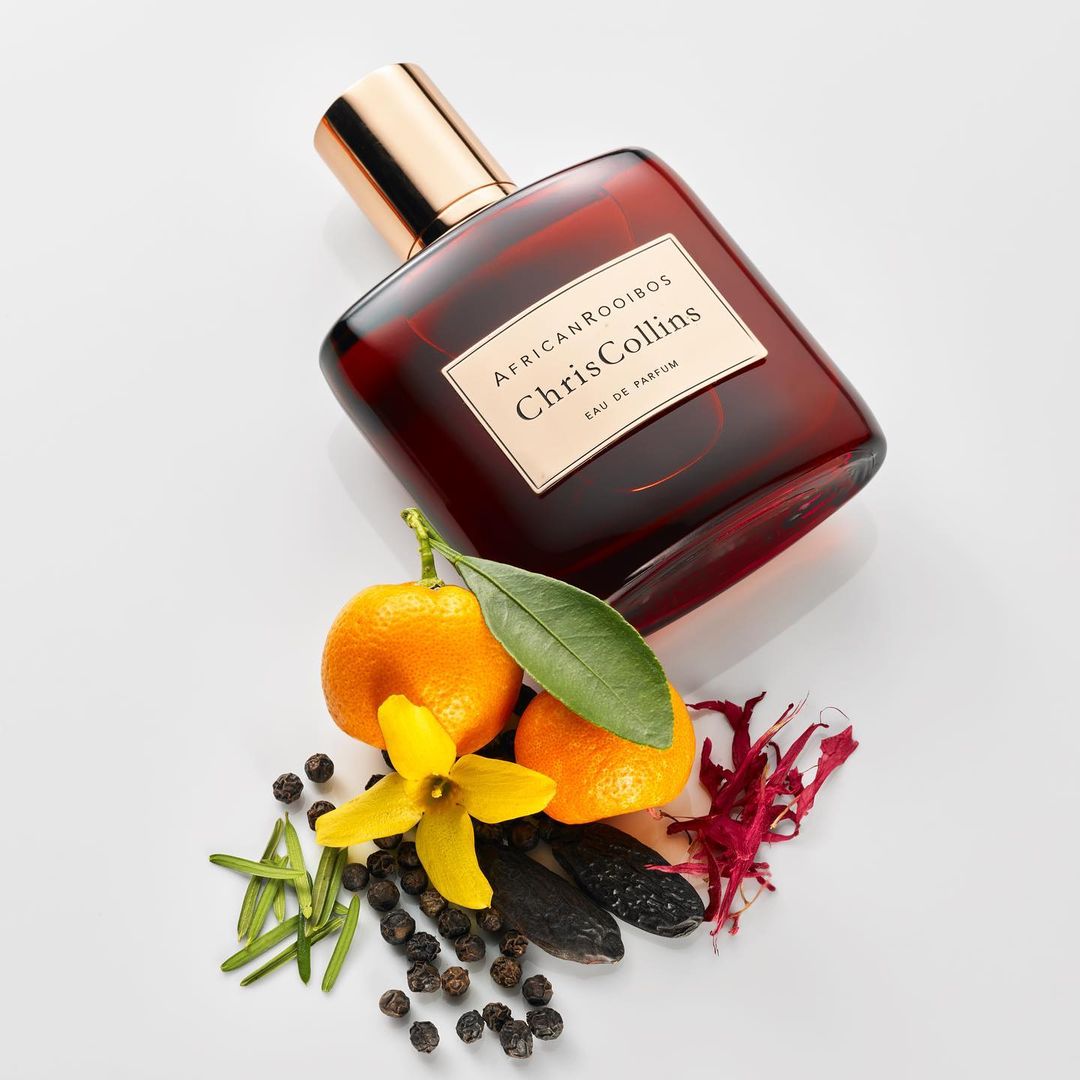 Overhead studio shot of amber glass perfume bottle with gold cap, tangerines, yellow flower, pepper, and spices on white background.