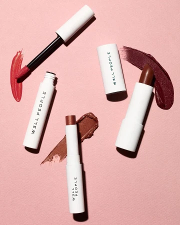 Flat lay of cosmetic products including lipsticks and liquid gloss tubes with matching makeup swatches on a pink textured background.