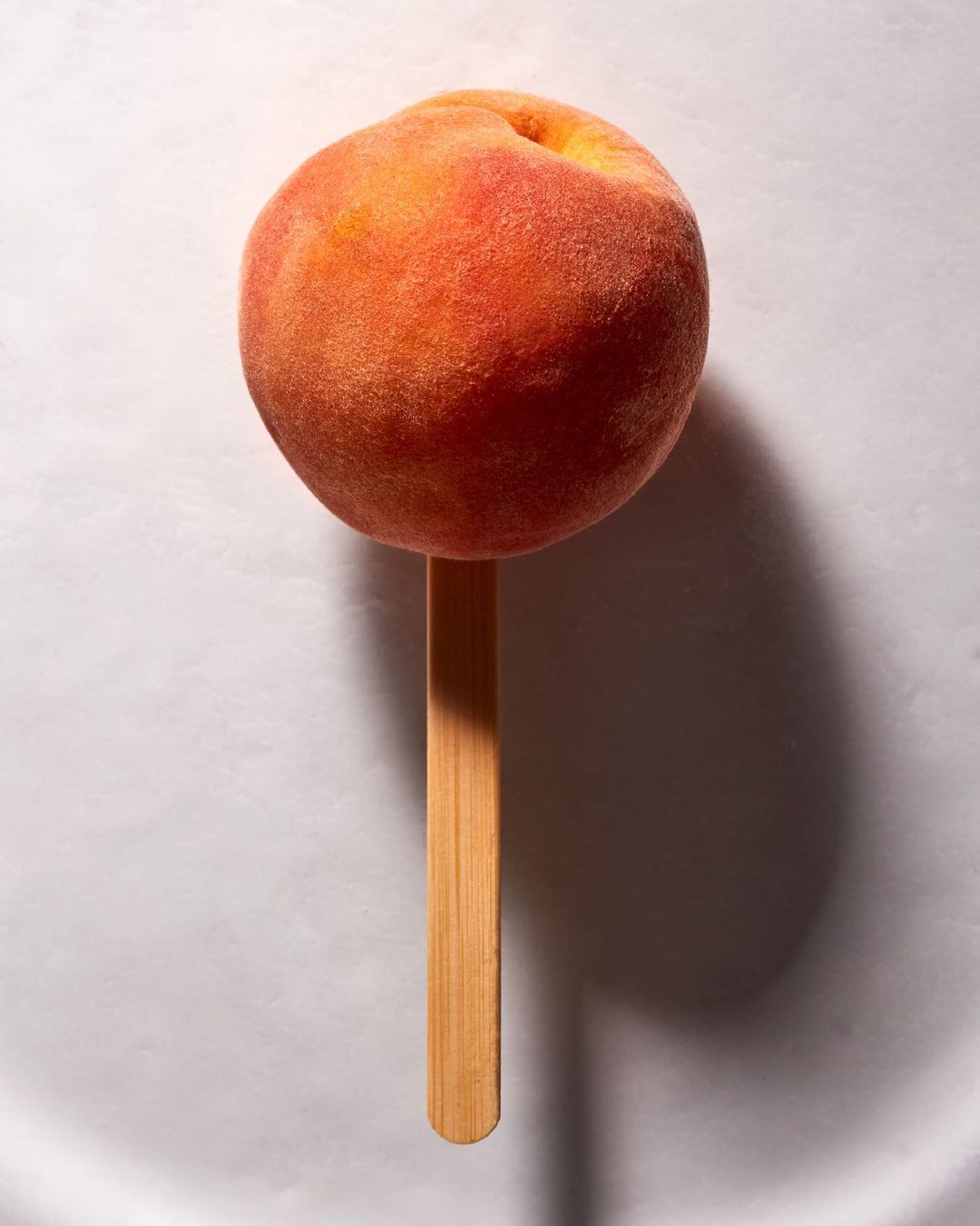 Single peach on a stick, set against a plain white background, showing the fruit's texture and coloration with dramatic lighting.
