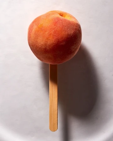 Single peach on a stick, set against a plain white background, showing the fruit's texture and coloration with dramatic lighting.