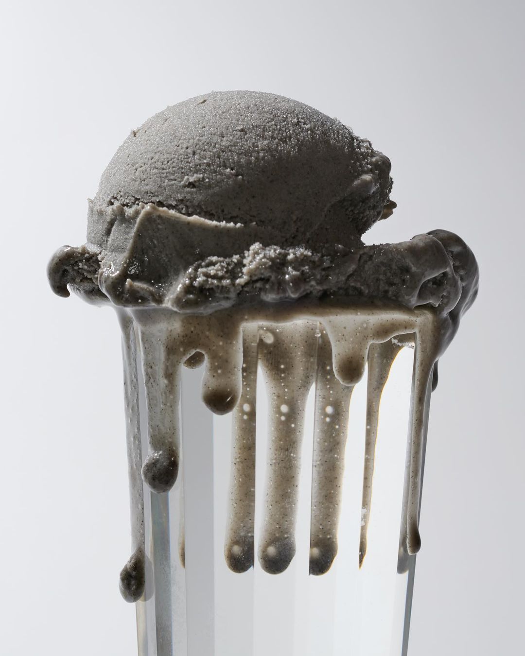 A scoop of grey ice cream melting down the side of a clear glass with visible condensation against a white backdrop. Food photography.