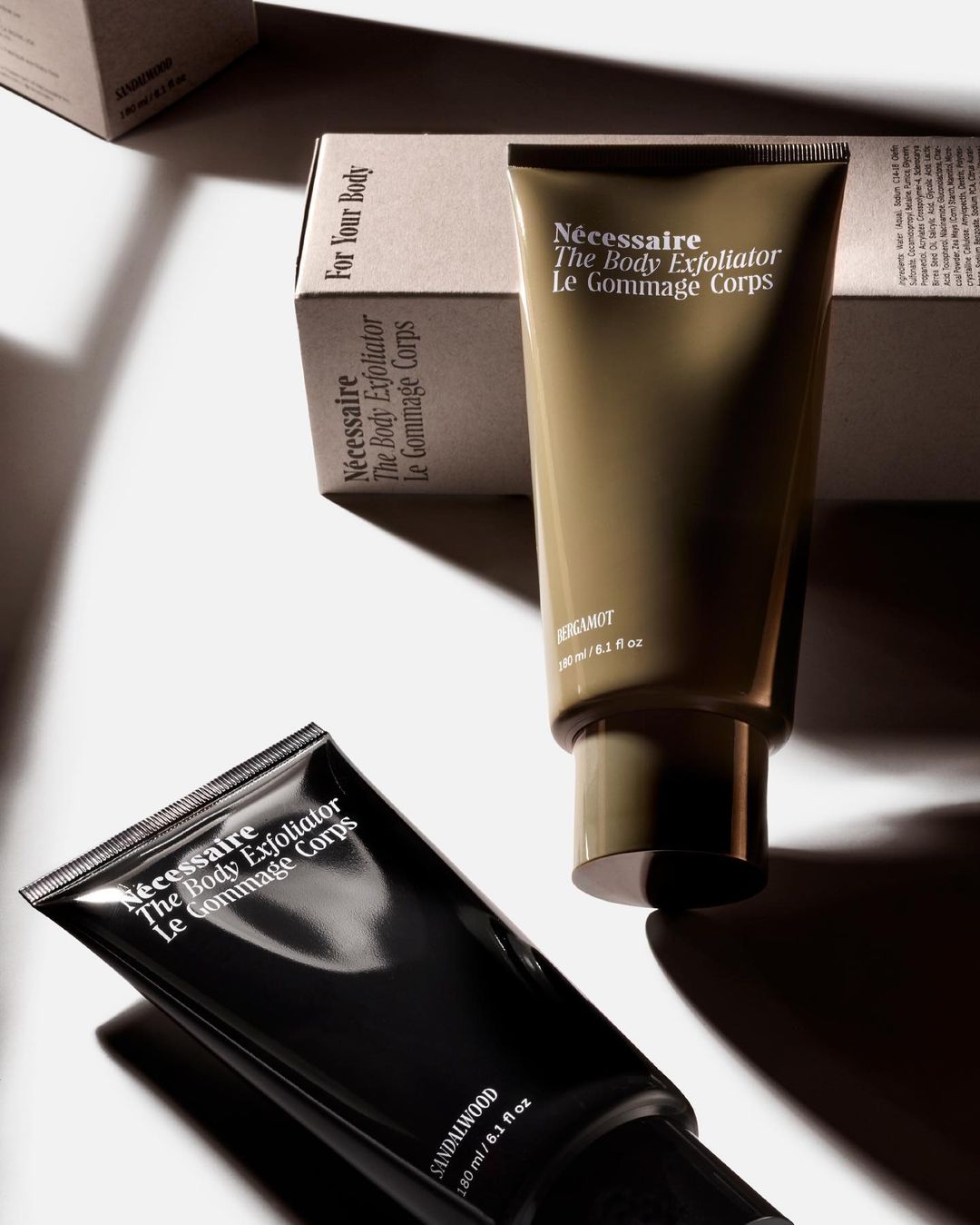 Two tubes of Nécessaire body exfoliator, one dark and one light, angled on a white surface with the product box in the background.