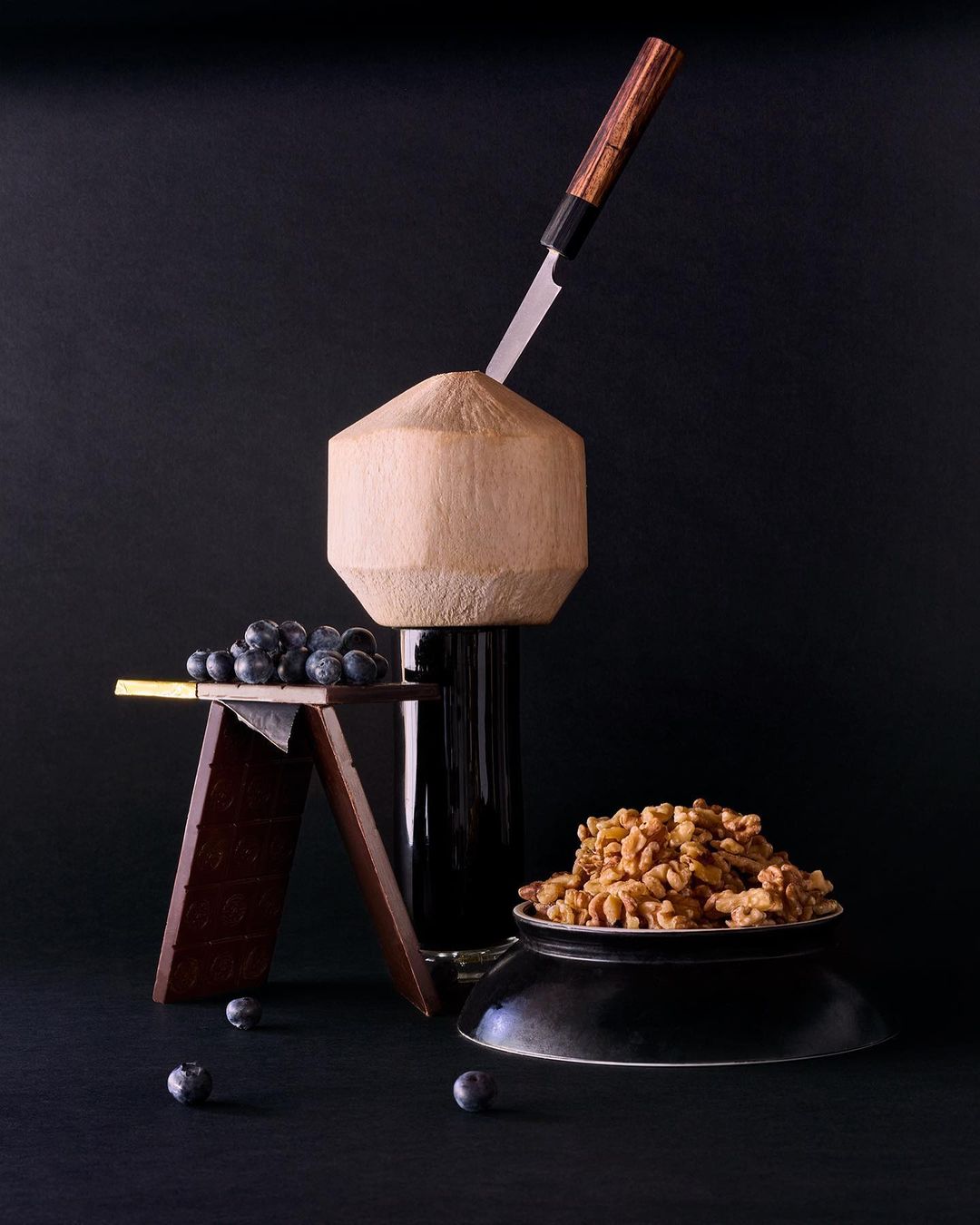 Still life featuring a halved coconut pierced by a knife, next to a wooden stand with blueberries and a bowl of walnuts against black backdrop.