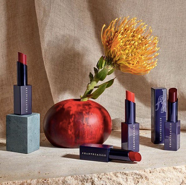 Cosmetic still life: Four Chantecaille lipsticks, one box, an orange flower in a vase, and a blue cube on a cream surface.