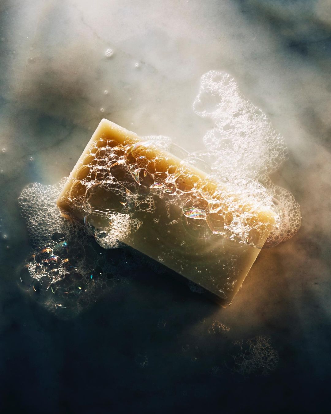 Close-up shot of a rectangular bar of soap surrounded by bubbles, set against a white and grey mottled surface.