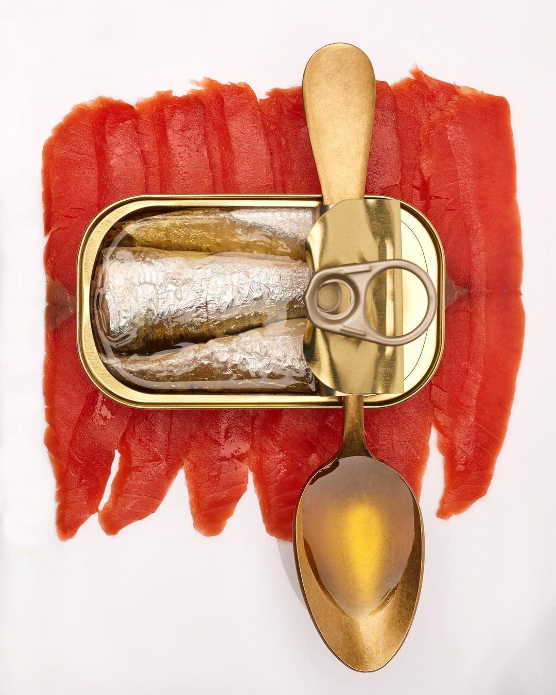 An opened metal can of sardines alongside a golden spoon and red fish slices, all arranged on a clean, white backdrop, overhead shot.