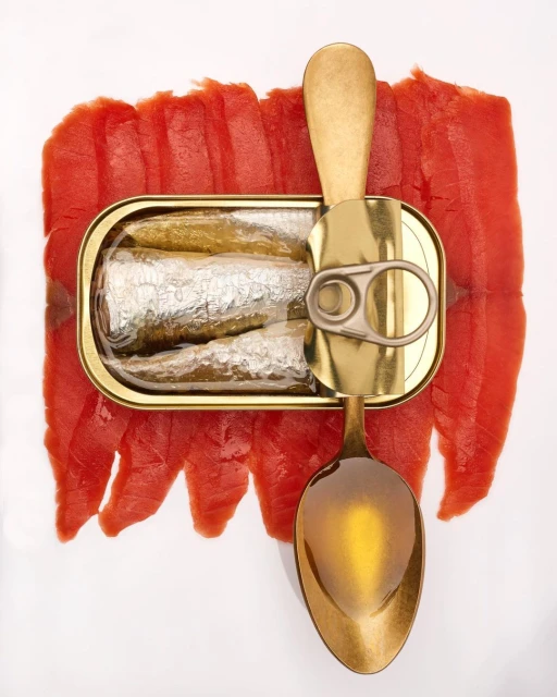 An opened metal can of sardines alongside a golden spoon and red fish slices, all arranged on a clean, white backdrop, overhead shot.