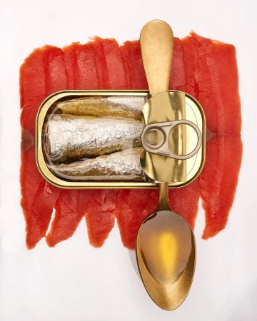 An opened metal can of sardines alongside a golden spoon and red fish slices, all arranged on a clean, white backdrop, overhead shot.