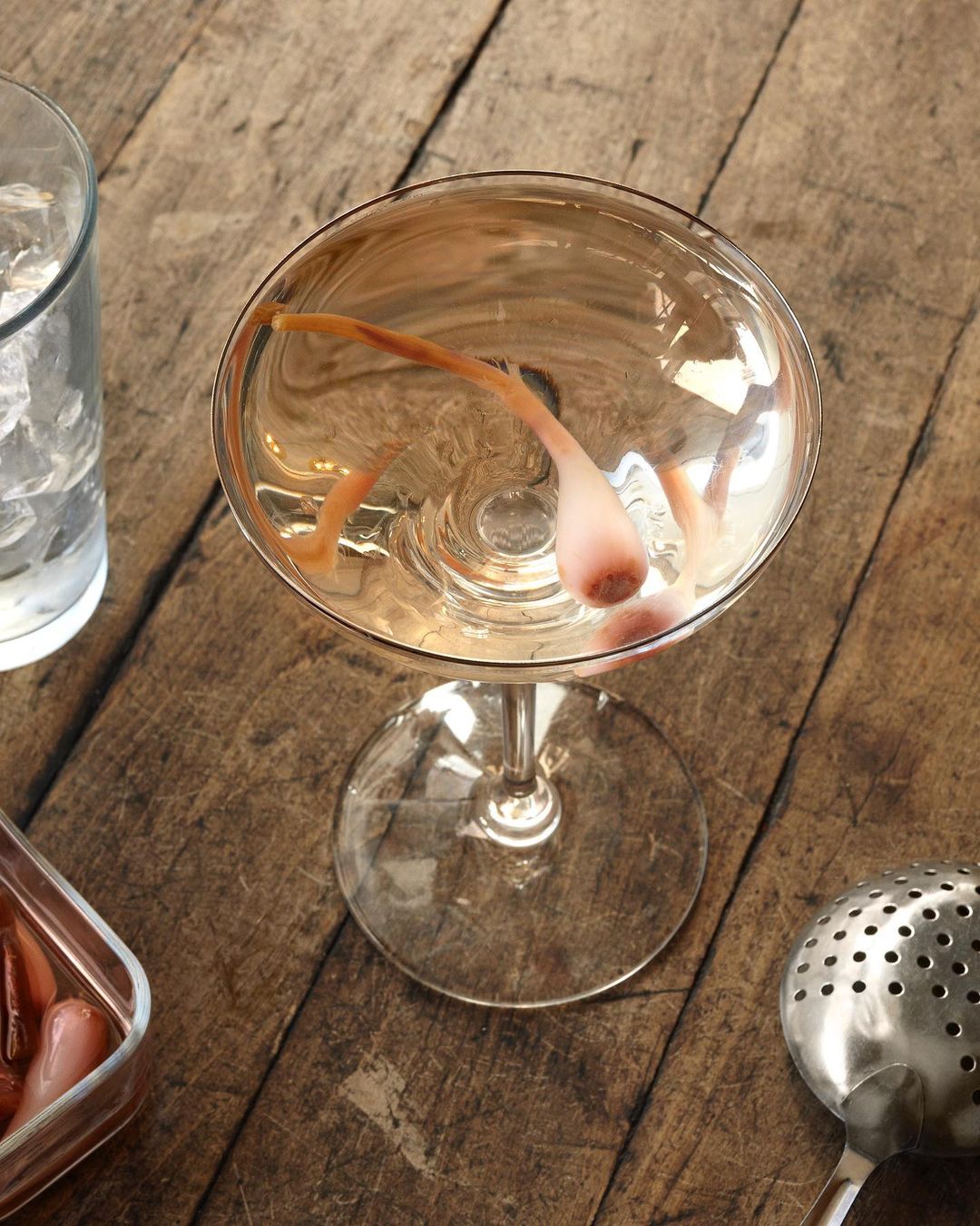 Overhead shot of a cocktail glass filled with a light-colored drink and a garnish, with a clear glass of ice and strainer nearby.