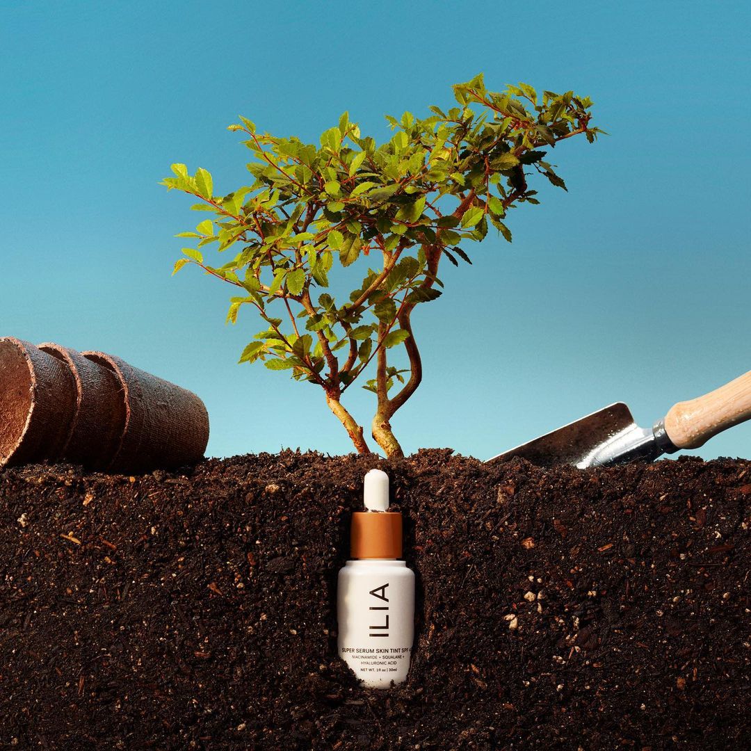 Cosmetic serum bottle nestled in soil, with a small green tree, gardening pots, and trowel on a bright blue background.