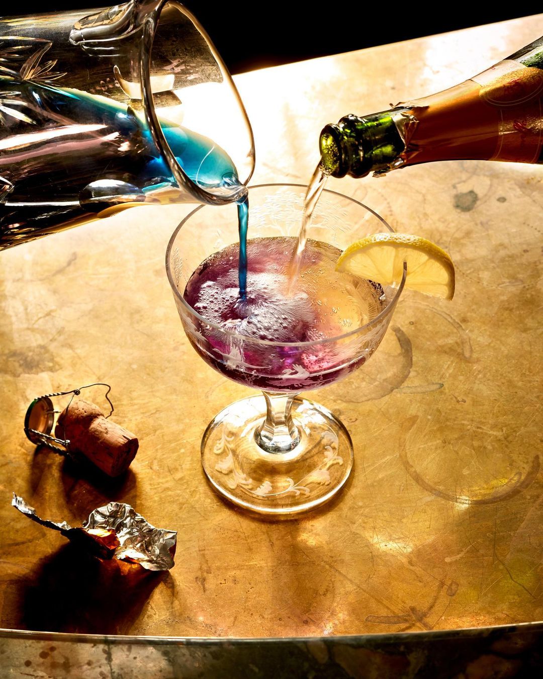 Two clear pitchers pour vibrant blue and clear liquid into a stemmed cocktail glass garnished with a lemon slice on a gold surface.