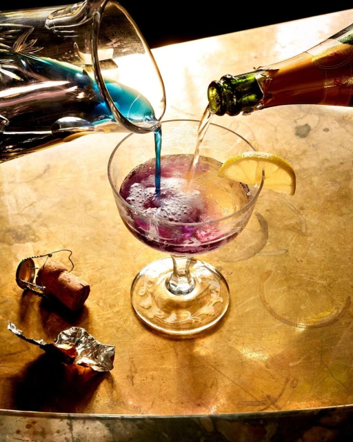 Two clear pitchers pour vibrant blue and clear liquid into a stemmed cocktail glass garnished with a lemon slice on a gold surface.