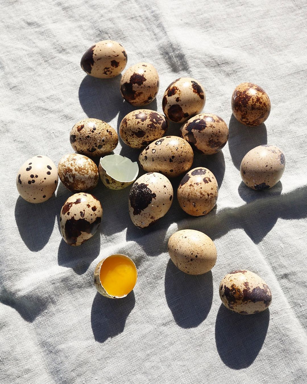 Overhead shot showcasing a group of quail eggs, some whole and some cracked open, arranged on a light green textured cloth surface.