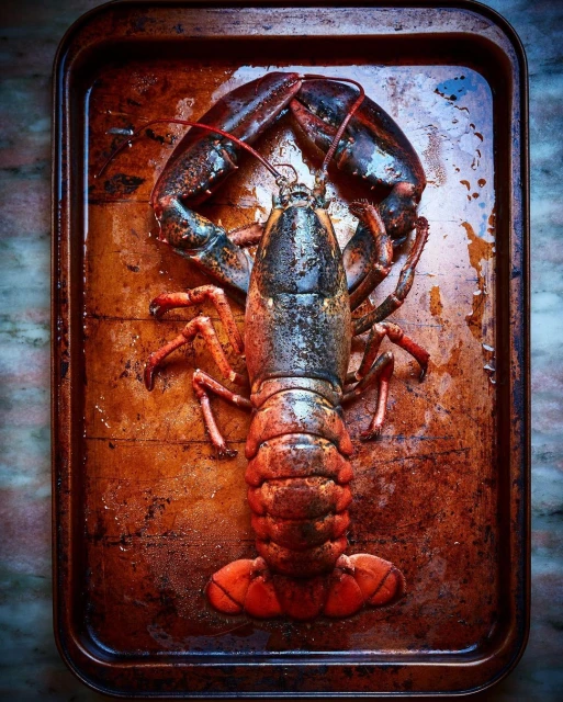 Overhead shot of a cooked lobster on a dark metal baking sheet with a distressed finish, in warm hues of red and brown.