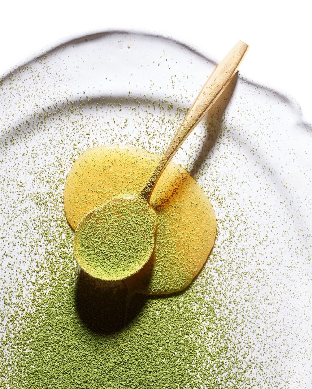 Wooden spoon filled with green matcha powder on a plate with honey and matcha powder around it, in a still life shot.