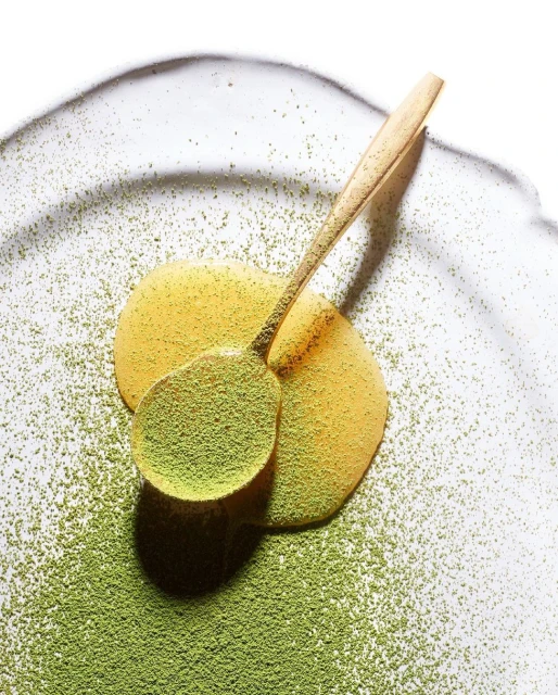 Wooden spoon filled with green matcha powder on a plate with honey and matcha powder around it, in a still life shot.