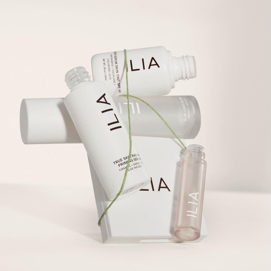 Cosmetic product bottles from Ilia including a serum and primer are stacked on a geometric block, connected by a green string.