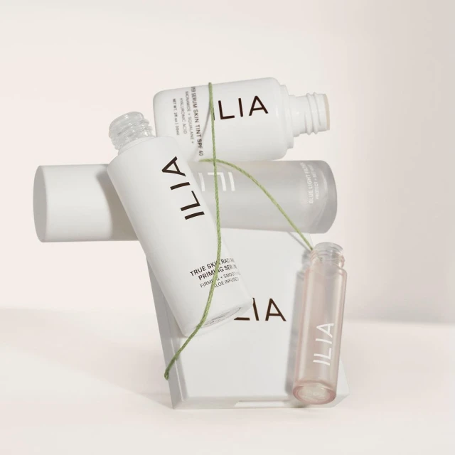 Cosmetic product bottles from Ilia including a serum and primer are stacked on a geometric block, connected by a green string.