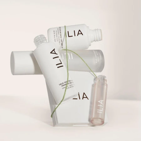 Cosmetic product bottles from Ilia including a serum and primer are stacked on a geometric block, connected by a green string.
