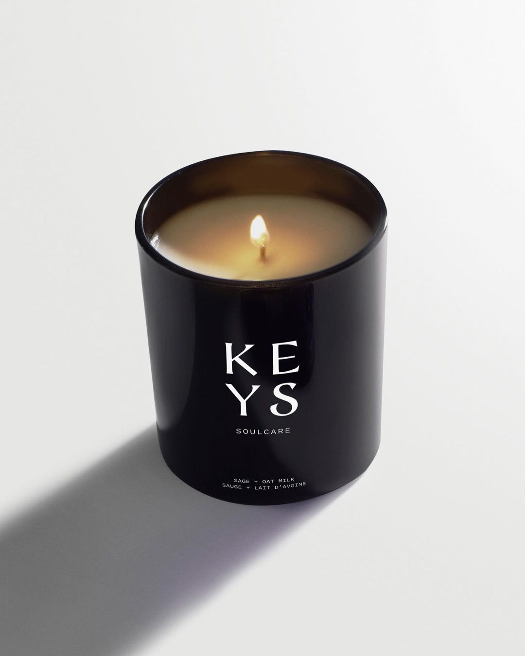 Minimalist studio shot of a black glass Keys Soulcare candle with a lit wick, casting a soft shadow on a white background.