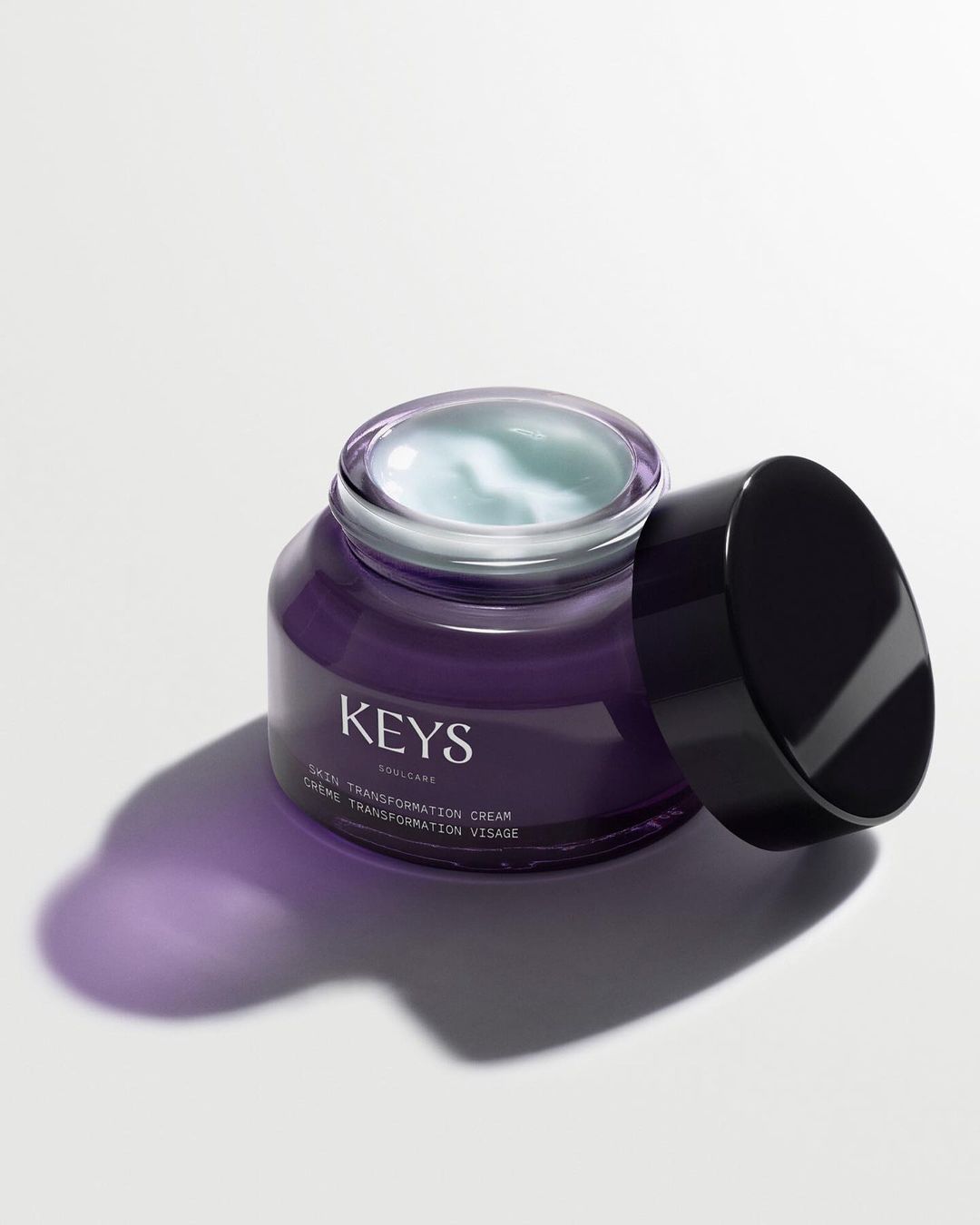 A container of skincare cream sits on a white surface, with a black lid placed next to the open jar, in a beauty product shot.