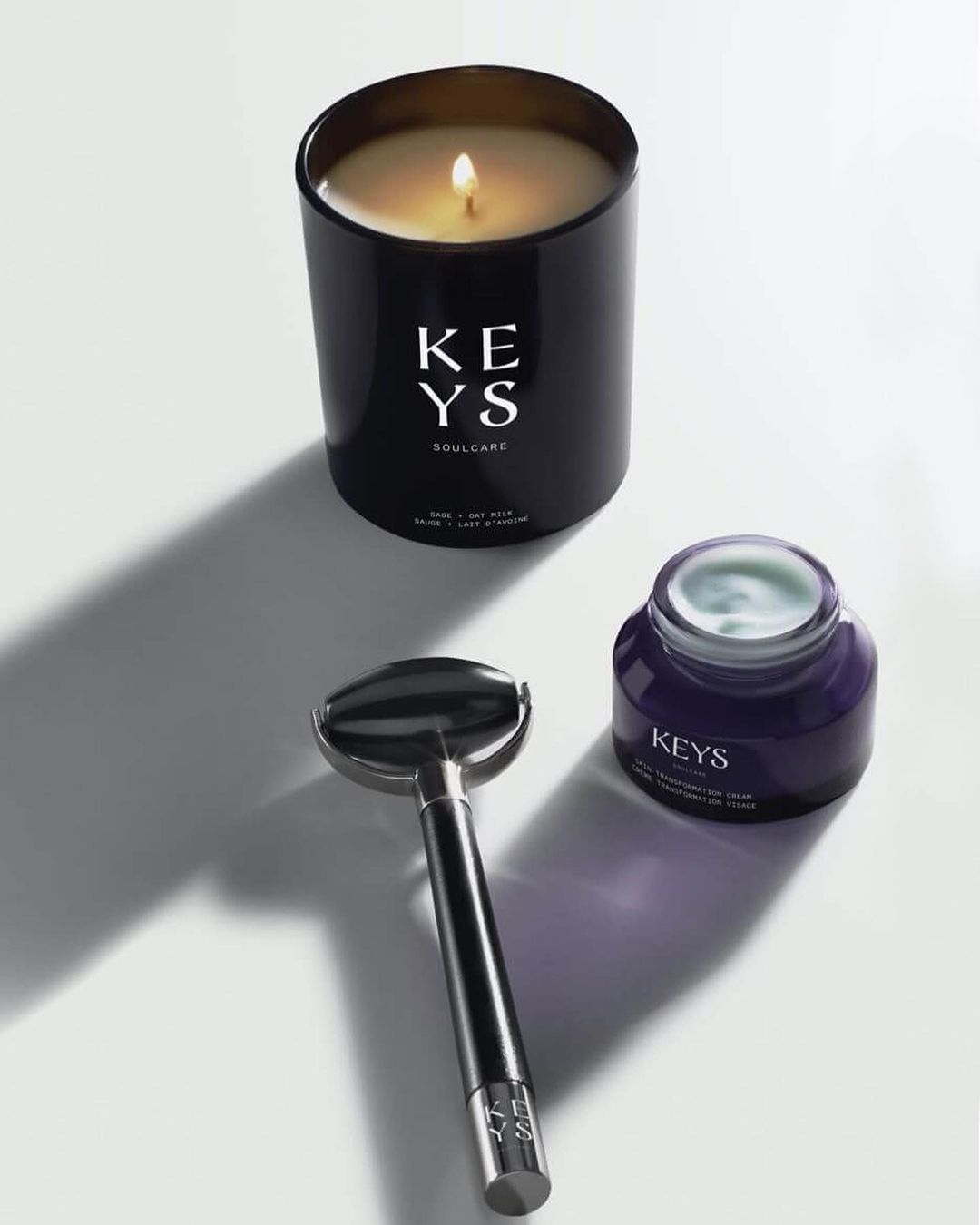 Still life studio shot of a black candle, silver face roller, and purple skincare jar on a white surface, showcasing beauty and wellness products.