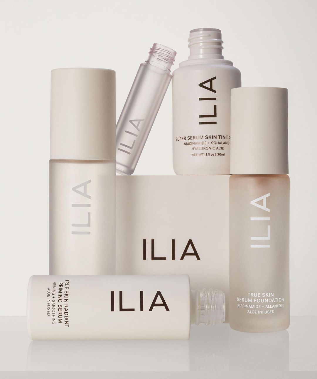 Several bottles and containers of Ilia beauty and skincare products are arranged together against a soft, neutral background.
